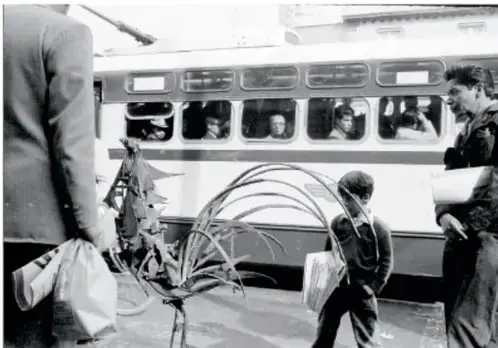 Fotografía en blanco y negro: un niño camina junto al gallo de hierro cerca de un camión o tranvía de pasajeros, mientras otros transeúntes lo observan.
