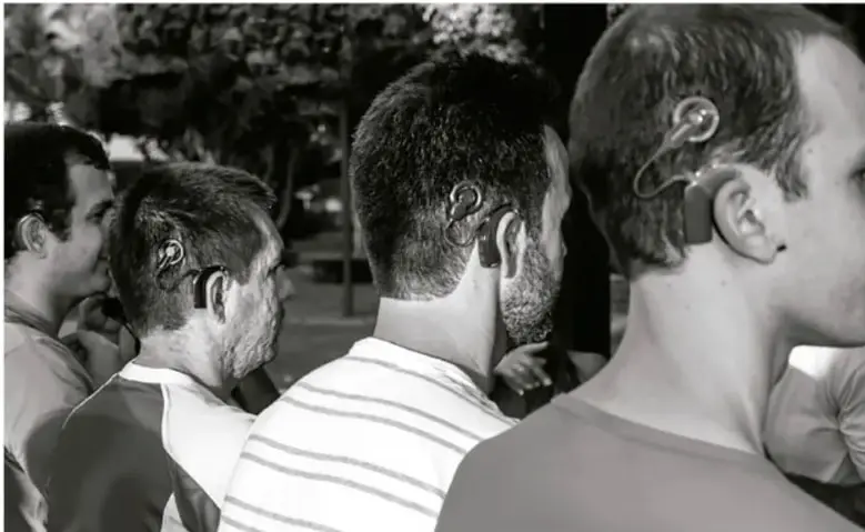 Fotografía en blanco y negro que muestra a varias personas de perfil con audífonos o dispositivos cocleares detrás de la oreja, en un espacio exterior.