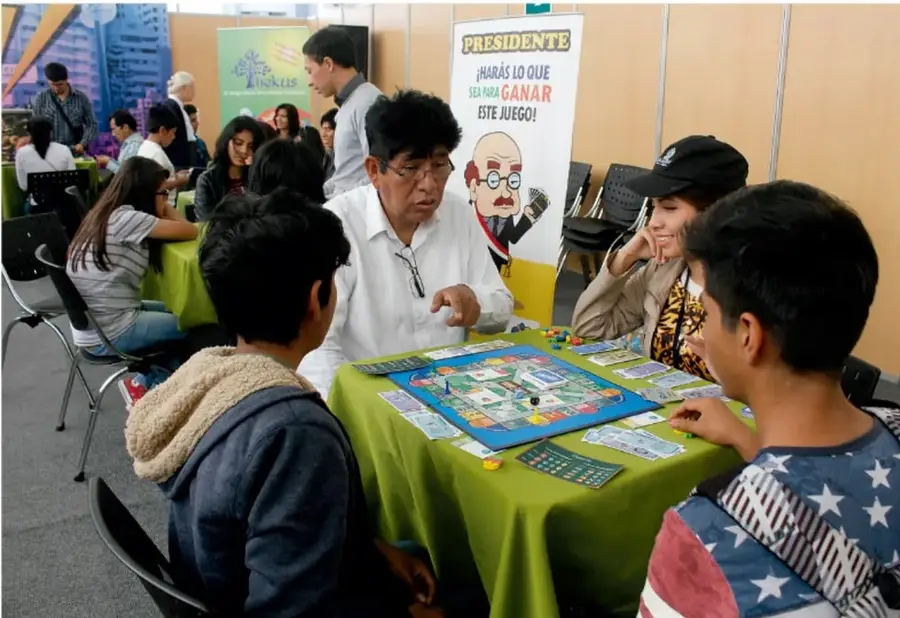 Fotografía de un grupo de personas, adultos y niños, jugando un juego de mesa en un tianguis lúdico. Se ve un cartel que dice 'PRESIDENTE' y otro que dice 'HARÁS LO QUE SEA PARA GANAR ESTE JUEGO'.
