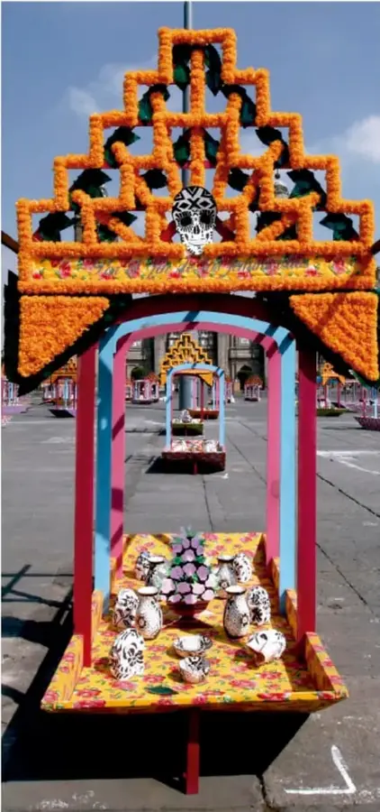 Ofrenda de Día de Muertos con arco de flores de cempasúchil, monos de calavera (calacas) y estructura ornamentada en tonos naranjas, rosas y azules, en una plaza pública.