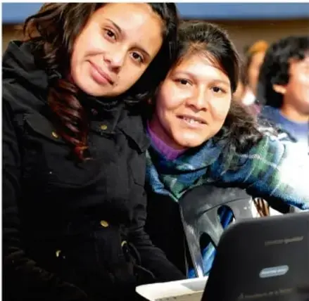Dos mujeres jóvenes sonríen frente a una computadora portátil, en un entorno educativo o laboral.