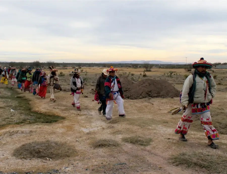Fotografía a sangre de una procesión de personas con vestimenta indígena tradicional (sombreros bordados, camisas y pantalones con motivos rojos) caminando por un camino de tierra en un paisaje árido al atardecer, con montículos de tierra y matorrales al fondo.
