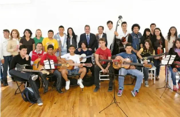 Fotografía de un grupo de estudiantes y docentes con instrumentos musicales (guitarras, teclado) en un salón con piso de madera, posando tras una actividad comunitaria.
