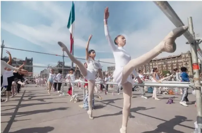 Fotografía de estudiantes de danza o ballet en un espacio público al aire libre, con una bandera de México al fondo y barras de práctica (barra de ballet) montadas en la explanada; muestra la puesta en práctica comunitaria de actividades recreativas.