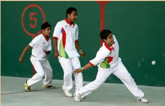 Fotografía de tres estudiantes adolescentes con uniforme blanco practicando frontón en una cancha techada; uno de ellos, en posición de golpe, mientras observa la jugada, y los otros dos se disponen a reaccionar. En la pared de la cancha se distingue la marca circular con el número 5.