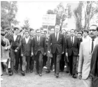 Fotografía en blanco y negro de una marcha estudiantil. Varias personas, principalmente hombres jóvenes, caminan por una calle. Al fondo se ve un cartel que dice 'ESTUDIANTES'. La imagen refleja una protesta pacífica.
