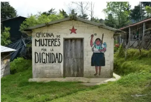 Fotografía de una pequeña construcción de concreto con techo de lámina, identificada con las palabras 'Oficina de Mujeres por la Dignidad' y una estrella roja. A un costado se observa un mural de una mujer con el puño en alto.