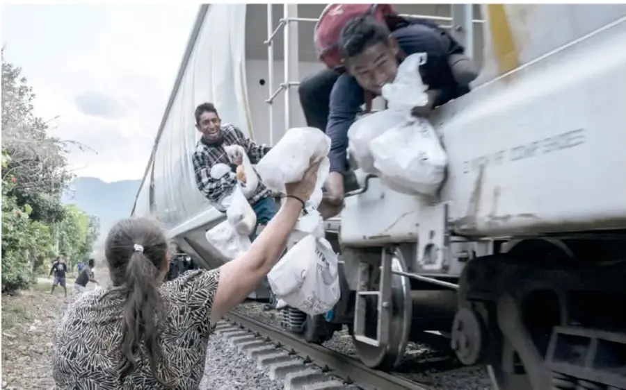 Fotografía de personas migrantes sirviéndose comida en platos y bolsas, sentadas sobre el pasto, durante una jornada de apoyo.