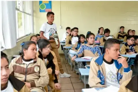 Fotografía de un grupo de estudiantes de secundaria en un aula; un alumno de pie participa mientras sus compañeros escuchan sentados en sus bancas.
