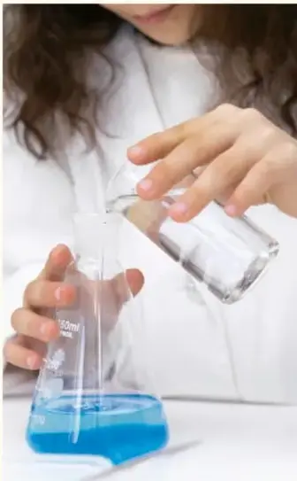 Estudiante vierte líquido azul de un vaso a un matraz Erlenmeyer, ilustrando el manejo de instrumentos de laboratorio.
