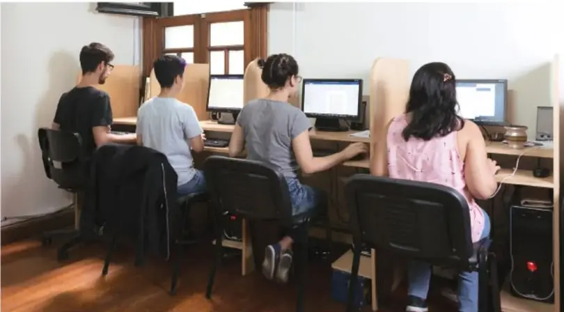 Fotografía de cuatro personas sentadas frente a computadoras de escritorio en una sala con paneles divisorios, probablemente un módulo de atención o centro de trabajo administrativo.