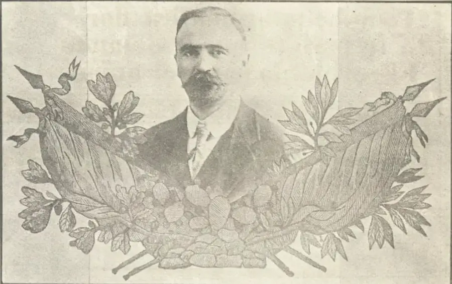 Retrato fotográfico de Francisco I. Madero, de medio cuerpo, con traje oscuro y corbata, enmarcado por ramas de laurel y olivo entrelazadas y cintas con trofeos en la base.