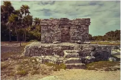 Fotografía de estructura de piedra en un sitio arqueológico con palmeras al fondo.