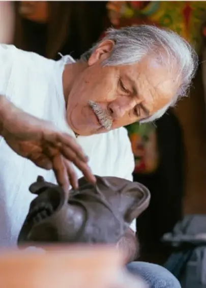Fotografía de un artista mayor con cabello blanco, modelando con las manos una pieza de barro o cerámica, en un taller artesanal.