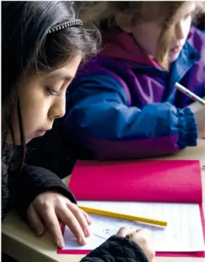 Fotografía de dos niñas trabajando en una mesa escolar; una de ellas escribe o dibuja sobre un cuaderno con un lápiz amarillo.