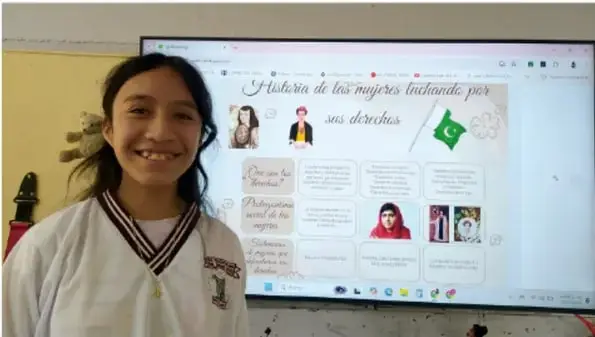 Fotografía de una estudiante sonriente frente a una pantalla que muestra un cartel digital titulado 'Historia de las mujeres luchando por sus derechos'.