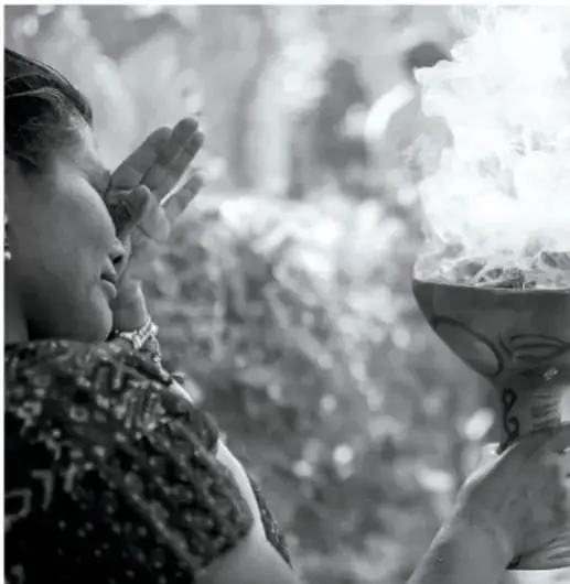 Fotografía en blanco y negro de una mujer en actitud de reverencia o rezando frente a un recipiente del que sale humo, probablemente quema de copal en un ritual.
