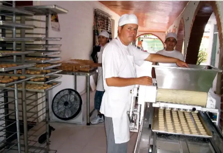 Fotografía de una panadería donde varios trabajadores con uniforme blanco elaboran pan; se observan charolas y una máquina para hacer pan.