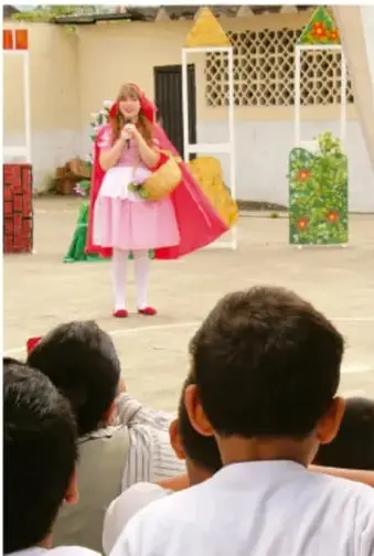 Fotografía de una niña caracterizada con capa roja representando un personaje en un escenario escolar, observada por un público de espaldas.