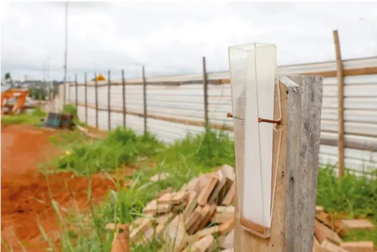 Fotografía de un pluviómetro instalado en un poste de madera, con un recipiente transparente graduado, en un entorno rural con vegetación y una cerca metálica al fondo.