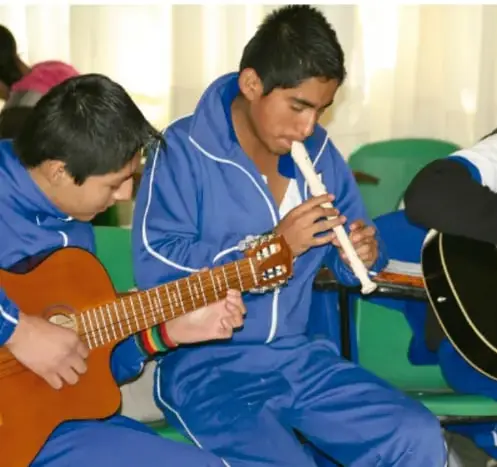 Fotografía de jóvenes estudiantes con uniforme azul; uno toca la guitarra y otro toca una flauta dulce (instrumentos musicales).