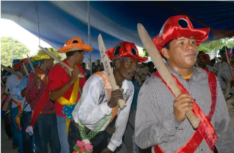 Fotografía de hombres de comunidades indígenas y afromexicanas participando en una celebración o danza tradicional, portando bastones de madera y sombreros de colores (rojo, naranja, amarillo) con cintas y accesorios ceremoniales, bajo una carpa azul.