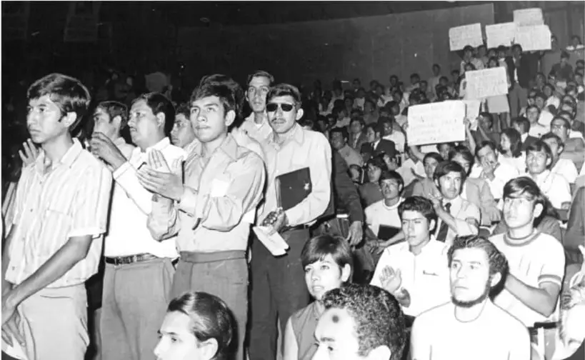 Fotografía en blanco y negro de una asamblea estudiantil numerosa, con jóvenes de pie y sentados, algunos con pancartas.