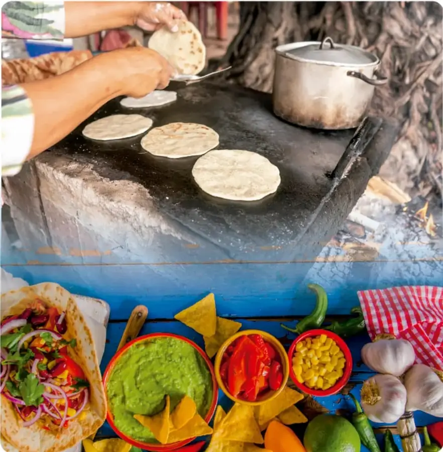 Fotografía en dos partes: arriba, manos preparando tortillas en un comal de metal sobre un fogón; abajo, una mesa con guacamole, totopos de maíz, maíz, ajo, limones, chiles, jitomate y una tortilla preparada.