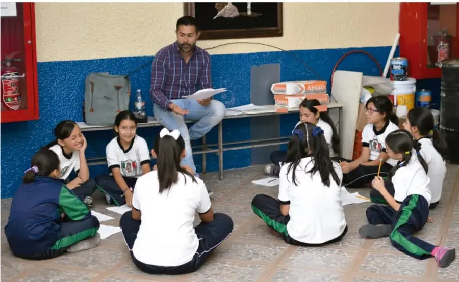Fotografía de un maestro sentado en un banco leyendo un libro frente a un grupo de estudiantes sentadas en el suelo formando un semicírculo, en un patio escolar con pared azul y extintor rojo.