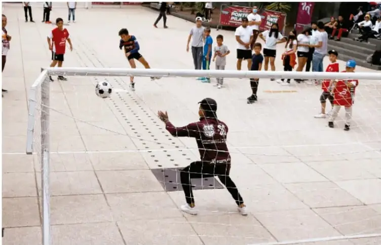 Fotografía de un partido de fútbol en una cancha urbana: un arquero se dispone a atajar un balón que se acerca a la portería, mientras otros jugadores y espectadores observan alrededor.
