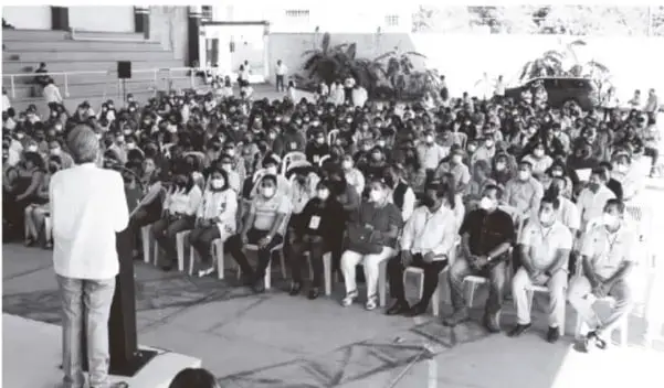 Fotografía en blanco y negro de una reunión masiva al aire libre: una persona de pie (visto de espaldas) frente a un amplio público sentado, aparentemente un acto o conferencia educativa.