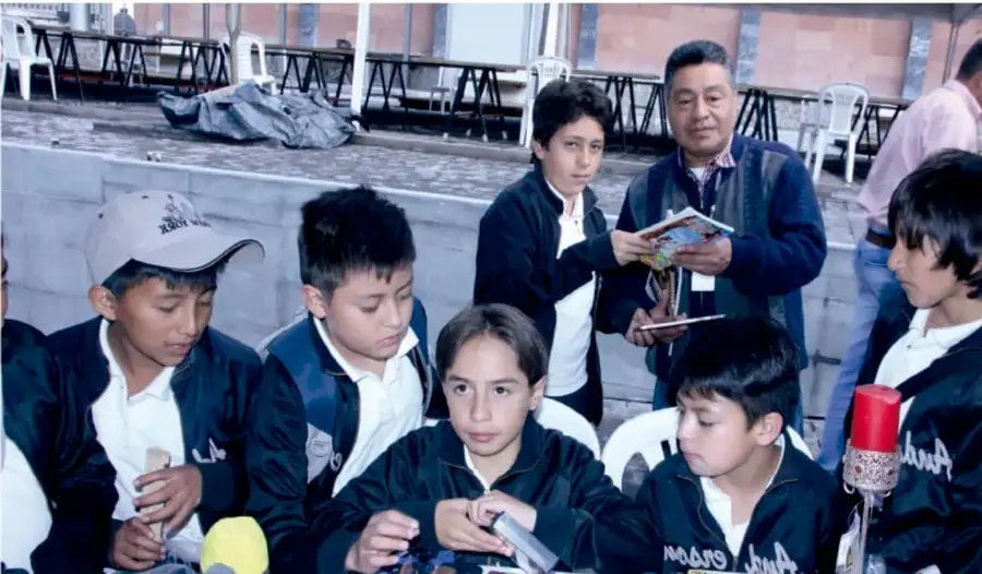 Grupo de estudiantes de primaria con uniforme escolar reunidos alrededor de una mesa, acompañados por un adulto que muestra un folleto; se observan materiales del puesto informativo.