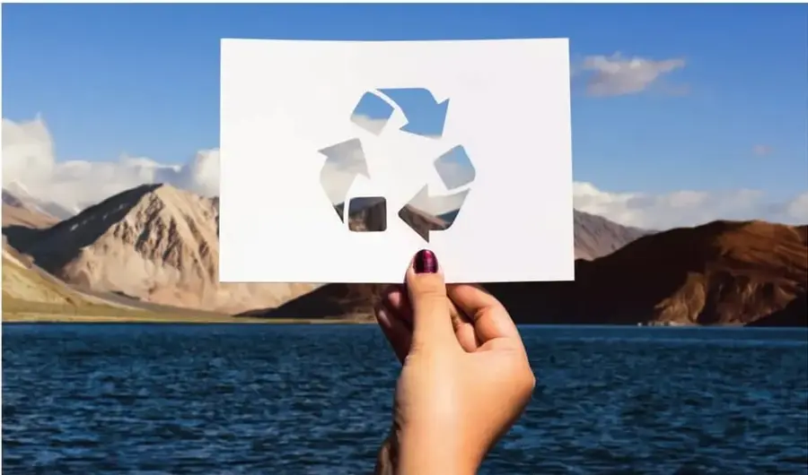 Foto realista de una mano sosteniendo un papel con el símbolo de reciclaje recortado, frente a un paisaje de lago y montañas.