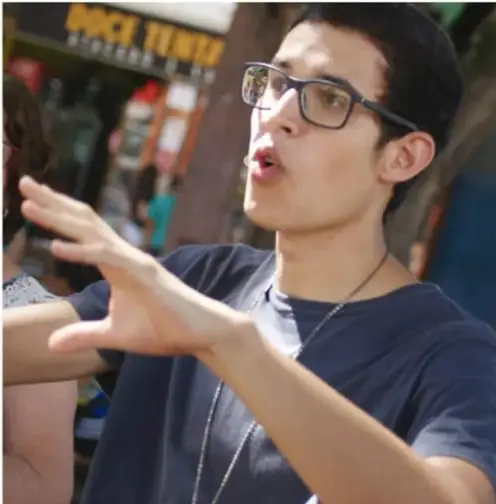 Fotografía de un joven estudiante con lentes, gesticulando mientras habla, probablemente presentando su guion o participando en la asamblea de retroalimentación.