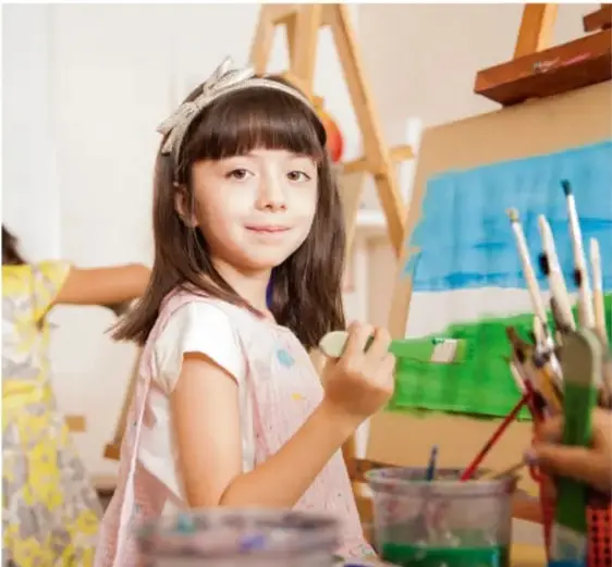 Niña con diadema sosteniendo un pincel frente a un caballete con pintura, en un salón de clases.