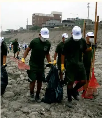 Fotografía de estudiantes con uniforme verde, guantes y bolsas, recogiendo basura en un terreno.