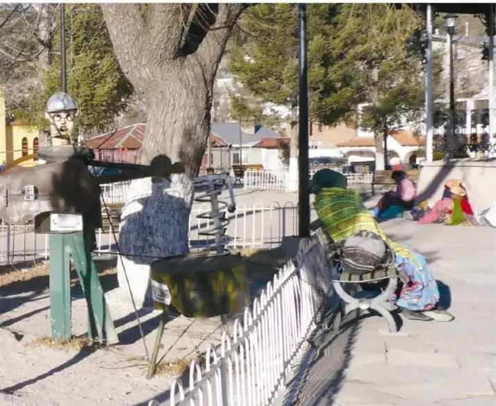 Plaza de un pueblo serrano: personas sentadas, algunas con vestimenta tradicional rarámuri de faldas de colores, vendiendo o descansando cerca de una banca, con árboles y bardas de colores.