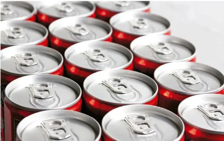 Fotografía de múltiples latas de refresco de aluminio vistas desde arriba, en tonos rojizos y plateados, ilustrando el consumo industrial de bebidas.