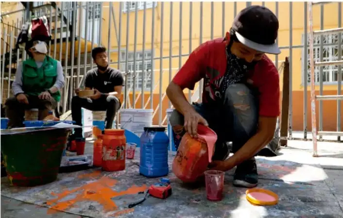 Fotografía de tres personas trabajando en la elaboración de un mural al aire libre. Una persona con gorra y camiseta roja sostiene una bolsa de pigmento frente a cubetas de pintura; otra persona con chaleco verde y cubrebocas observa sentada, y una tercera persona sentada toma notas. El piso está manchado de pintura naranja y hay andamios y rejas de fondo.