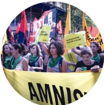 Fotografía de mujeres manifestantes con banderas y pancartas, sosteniendo una manta parcialmente visible que dice 'AMNIS' (Amnistía Internacional).