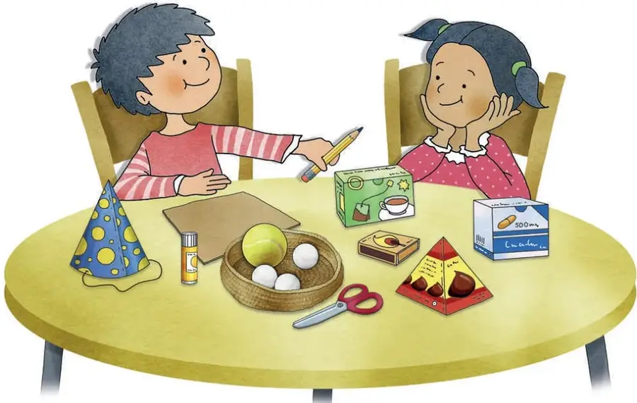 Dos estudiantes sentados a una mesa redonda con materiales reutilizables: un gorro de fiesta cónico azul con lunares, un cartón rectangular, una lata de spray, un canasto con pelotas grandes y pequeñas, una caja de leche, una caja triangular de chocolates, tijeras y un lápiz.