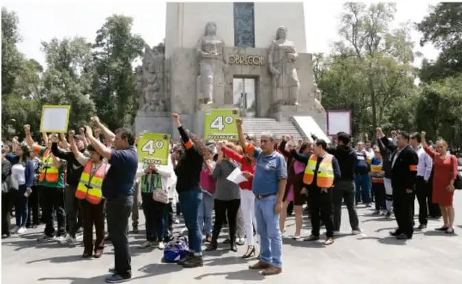 Grupo de personas reunidas frente a un monumento con pancartas que dicen '4.º' y los brazos levantados, participando en una actividad comunitaria.