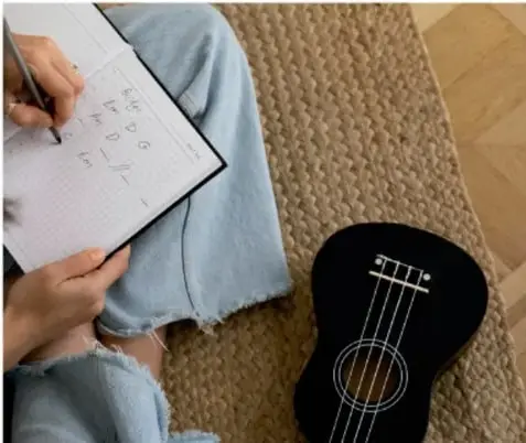 Fotografía de una persona sentada en el piso escribiendo en una libreta cuadriculada, con una guitarra negra de dibujo al lado.