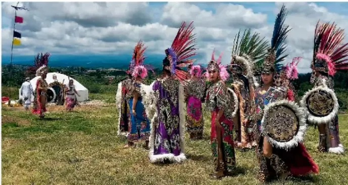 Fotografía de personas indígenas con vestimenta ritual colorida y penachos de plumas, en un entorno natural abierto con montañas al fondo, realizando una práctica ceremonial colectiva.