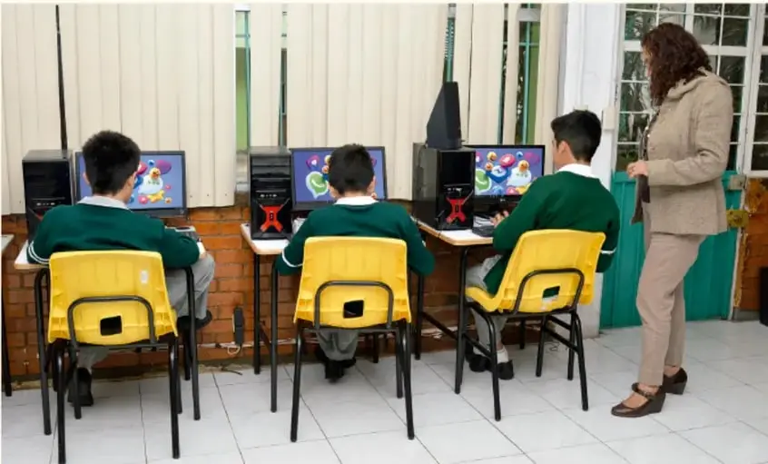 Fotografía de un aula de computación: tres estudiantes con uniforme verde sentados frente a computadoras de escritorio y una docente de pie supervisándolos.