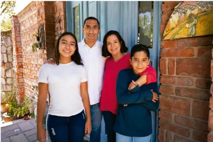 Fotografía de una familia de cuatro personas (dos adultos y dos jóvenes) sonriendo frente a la entrada de una casa de ladrillo, ilustrando la convivencia familiar.