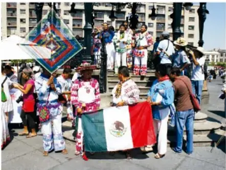 Personas con vestimenta tradicional y una bandera de México reunidas en una plaza pública.