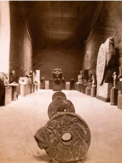 Fotografía en blanco y negro de una sala de museo con esculturas prehispánicas, entre ellas una piedra circular tallada en primer plano y figuras de piedra al fondo.