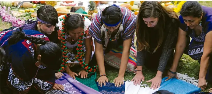Fotografía de un grupo de personas —jóvenes y adultas, varias con textiles y accesorios tradicionales— arrodilladas o agachadas sobre un suelo con mantas de colores, en lo que parece una actividad comunitaria o ceremonia al aire libre con frutas y ofrendas al fondo.