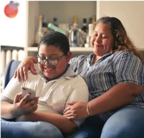Fotografía de una madre y su hijo sentados en un sofá; el niño usa un teléfono celular mientras la madre lo acompaña, ejemplificando el sedentarismo y el estilo de vida relacionado con la obesidad infantil.
